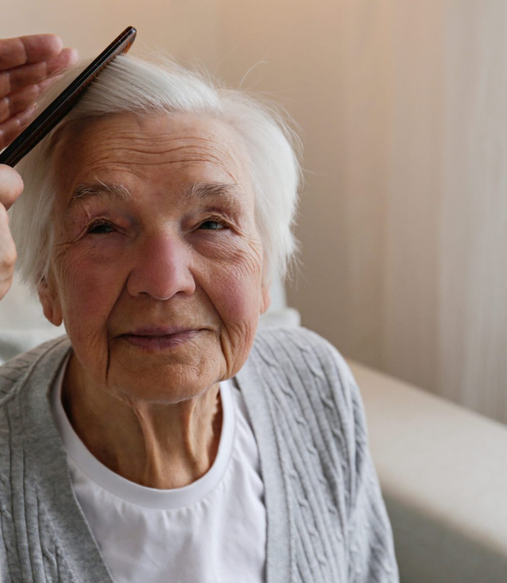 Unrecognizable female expressing care towards an elderly lady, brushing her hair with a comb. Granddaughter helping granny with a haircut. Family values concept. lose up, copy space, background.